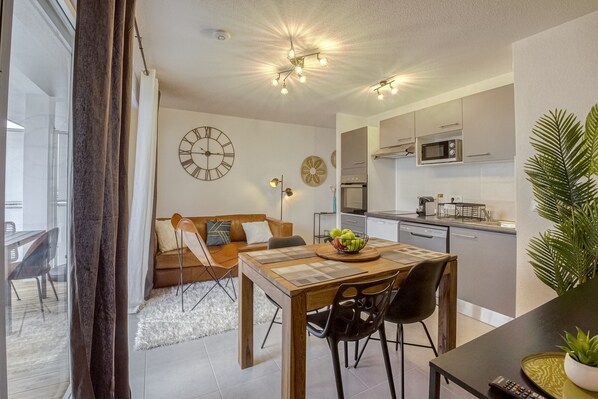Cocina moderna con microondas, horno y lavamanos. Hay un comedor con mesa y sillas de madera, y un sofá con cojines. Un reloj está en la pared y una planta en la encimera.