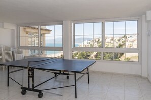 A spacious room with a ping pong table, large windows with a coastal view, and a tiled floor.