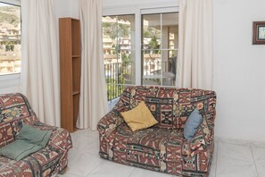 A living room with patterned sofas, a wooden bookshelf, and a balcony with a view of buildings.