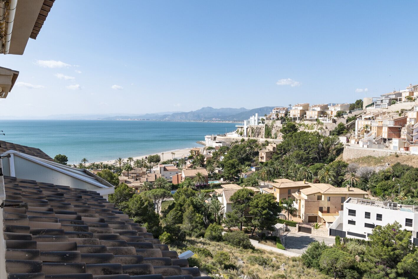 A coastal town with terracotta-roofed buildings, a beach, and a view of the sea and distant mountains.
