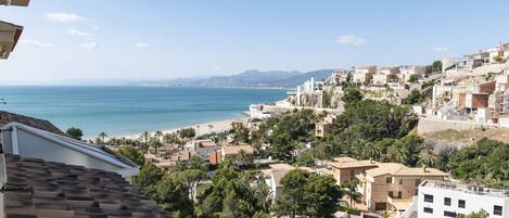 A coastal town with terracotta-roofed buildings, a beach, and a view of the sea and distant mountains.