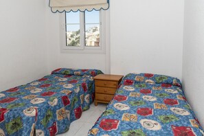 A bedroom with two single beds, a wooden dresser, and a window with a view of buildings.