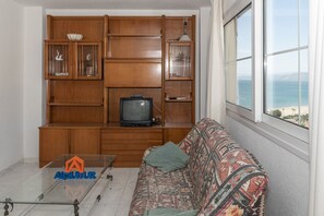 Una sala de estar con un mueble de entretenimiento de madera, un televisor, un sofá estampado y vista al océano a través de un ventanal grande.
