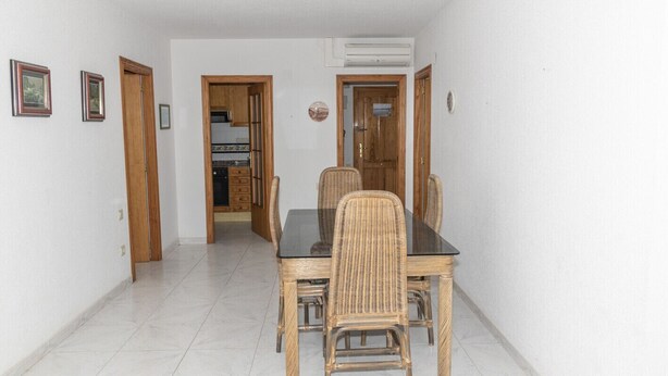 A dining area with a black table and wicker chairs, white tiled floor, and wooden doors leading to other rooms.