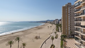 A beachside view with a sandy beach, palm trees, and a high-rise building with balconies. - EL RACO, A-7º-13 (Cullera)
