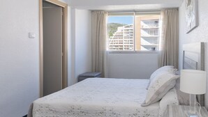 A hotel room with a bed, a nightstand, a lamp, a window with curtains, and a view of buildings outside. - EL RACO, B-8º-15 (Cullera)