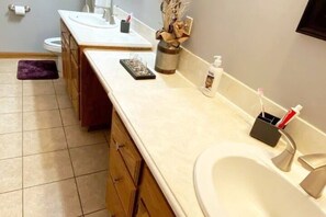 A bathroom with two sinks, a toilet, and a mirror. There are toiletries on the counter and a towel rack.