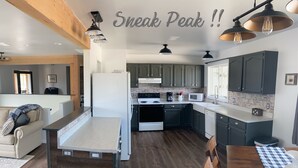 A modern kitchen with dark wooden flooring, a central island, and dark green cabinets. There is a dining table with a checkered tablecloth and a chair. - 20+ Acres of Peace @ The Saratoga Hoemstead (Encampment)