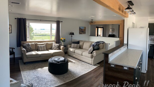 A spacious living room with a large window, a sofa set, a black ottoman, and a kitchen area with a refrigerator. - 20+ Acres of Peace @ The Saratoga Hoemstead (Encampment)