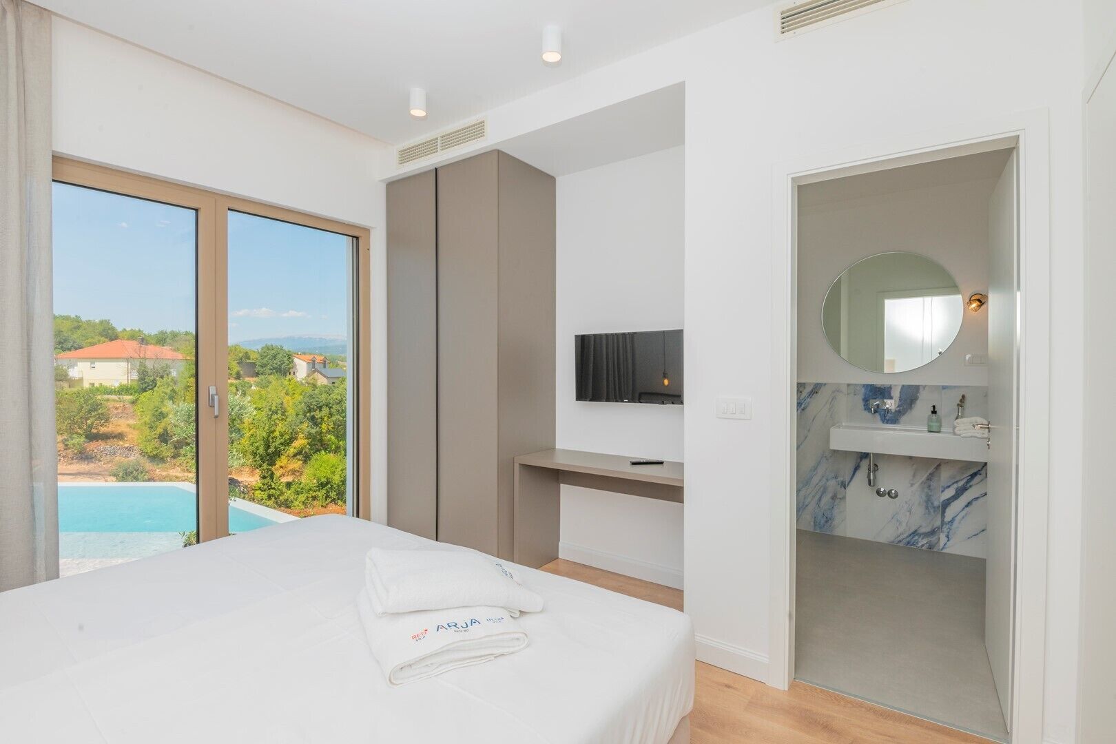 A hotel room with a bed, a TV, a bathroom with a marble sink, and a view of a swimming pool and greenery.