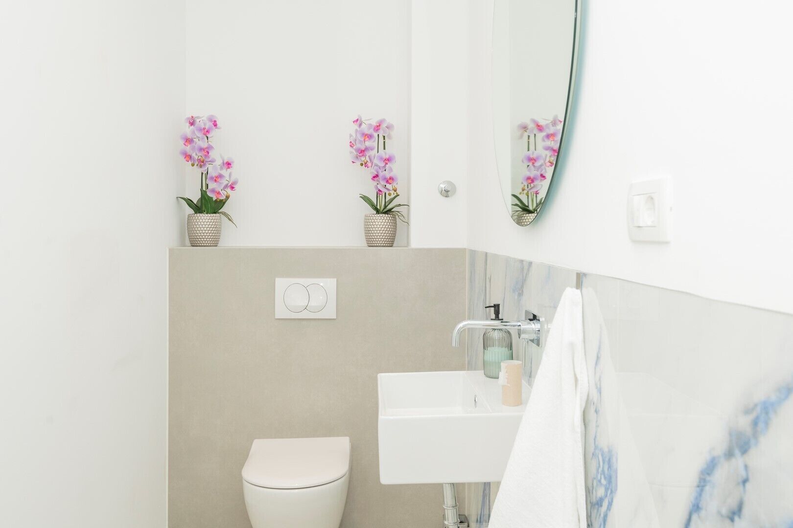 Ein modernes Badezimmer mit Toilette, Waschbecken und Spiegel. Über der Toilette befinden sich zwei Orchideen in Töpfen an der Wand.