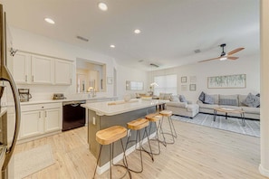 A modern kitchen with white cabinets, a central island with stools, a dining area with a sofa and coffee table, and a ceiling fan.