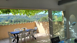 A balcony with a table and chairs, a sliding glass door, and a view of a forested hillside. - Gîte clim garage, Les Vans Ardèche, large panoramic terrace, gorges Ardèche (Les Assions)