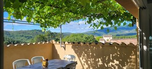 A balcony with a table and chairs, a view of green hills, and a building in the distance.