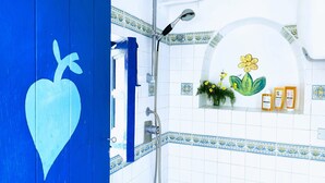 Une salle de bain avec une porte bleue, des murs recouverts de carreaux blancs, une douche et une étagère où sont posés des articles de toilette et un pot de fleurs.