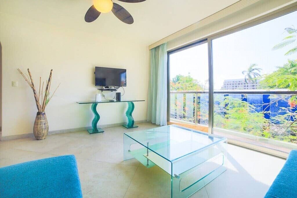 Ein modernes Wohnzimmer mit einem Glastisch, einem Flachbildfernseher auf einem Lowboard, einer Vase mit Trockenpflanzen und einem Balkon mit Blick auf Grünflächen und Gebäude.