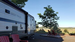 Un camping-car WILP gris est garé, avec une terrasse et deux chaises Adirondack placées face à un arbre et à un champ dégagé.