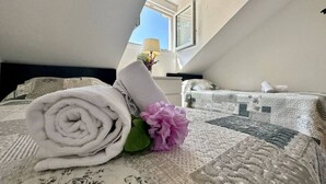 Un lit bien fait, avec une serviette blanche roulée et une fleur rose, une fenêtre donnant sur le ciel et une lampe au plafond.