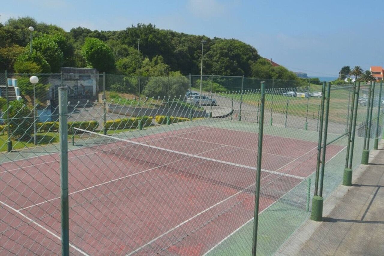 Un court de tennis entouré d’une clôture en treillis métallique, bordé de verdure et doté d’un espace de stationnement.