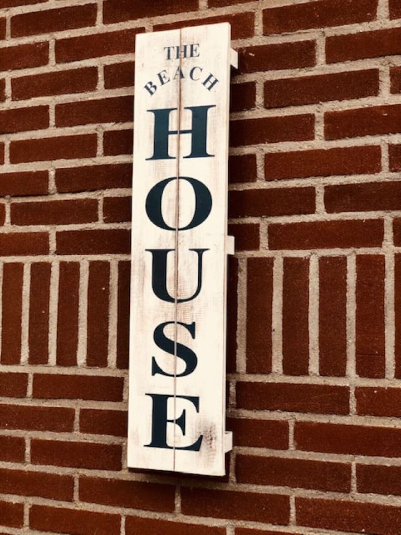 A wooden sign with "THE BEACH HOUSE" written on it, mounted on a brick wall.