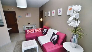 A living room with a red sofa, white coffee table, and a small white table. There are framed pictures on the wall and a vase with a plant on a small table.