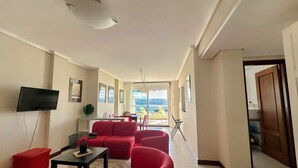 A spacious living area with a red sofa set, a flat-screen TV mounted on the wall, and a view of the sea through large windows.
