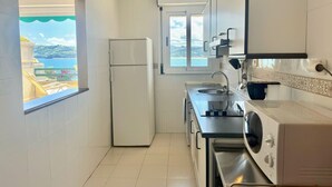 A compact kitchen with a refrigerator, sink, and washing machine. A window offers a view of the sea.