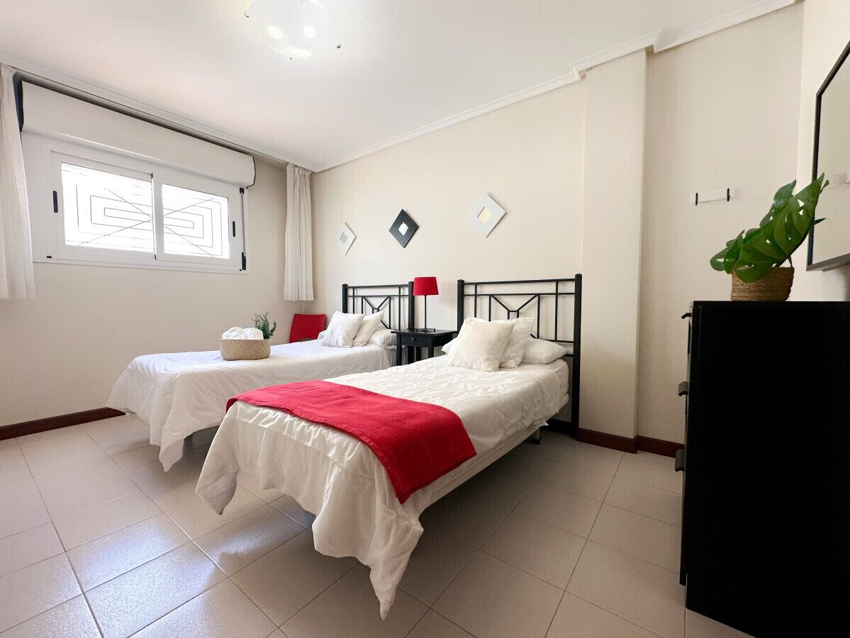 Habitación de hotel con dos camas, una mesita con una cesta, una lámpara roja y una planta en maceta.