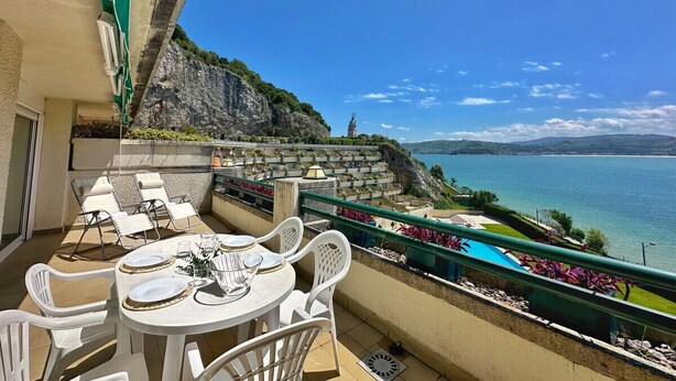 A balcony with a table set for dining, overlooking a coastal landscape with a pool and a rocky cliff.