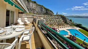 A terrace with white chairs and a table set for dining, overlooking a swimming pool and a scenic coastal view.