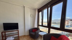A room with a TV on a stand, two armchairs, a small round table, and large windows with a view of a body of water and buildings.