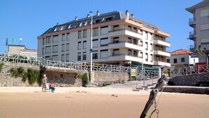 A multi-story building with balconies, a sandy beach in the foreground, and a stone wall.