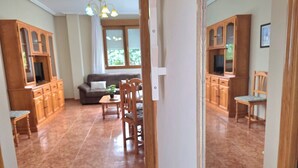 A spacious living room with a sofa, dining table, and chairs. There are wooden cabinets and a window with a view.