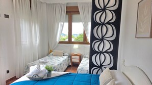 A bedroom with a large bed, a smaller bed, a nightstand, a lamp, and a window with curtains.