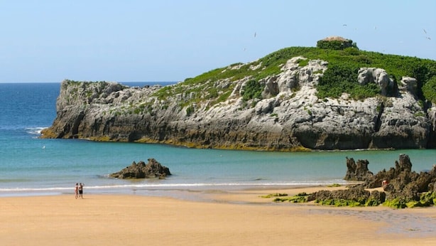 A beach with rocky cliffs, clear blue water, and a few people on the sand.