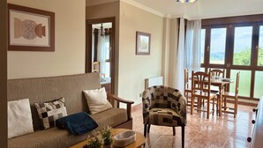 Un salon comprenant un canapé, un fauteuil, une table à manger et des chaises. Une fenêtre offre une vue sur la verdure à l’extérieur.