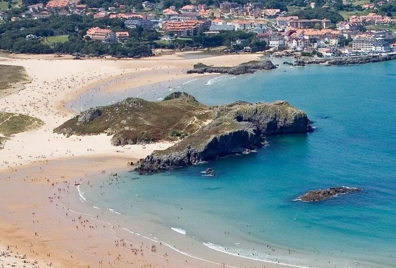 A coastal town with a beach, rocky outcrops, and buildings in the background.