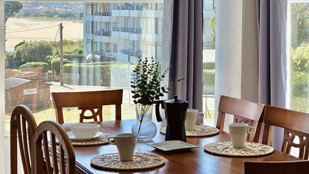 Un comedor con una mesa lista para cuatro personas, una cafetera y un jarrón con hojas verdes. La habitación tiene ventanas amplias con cortinas, que ofrecen vistas a un edificio y una playa.