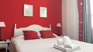 Habitación de hotel con pared roja, cama blanca, almohadas rojas y cabecero blanco. Hay dos mesitas de noche con lámparas y una ventana con cortinas.
