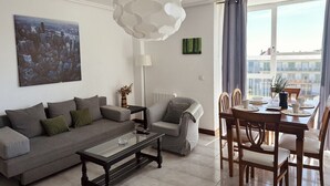 Una sala de estar moderna con un sofá, una mesa de centro y una mesa de comedor preparada para cuatro. Un cuadro de paisaje urbano y una obra de arte abstracta verde decoran las paredes.