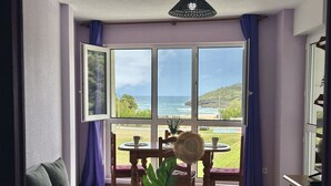 A room with a large window, a dining table with chairs, a sofa, and a small table with a vase and a hat. - Apto en primera línea - La Arena 4107 (Arnuero)