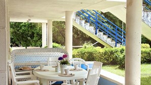 A covered patio with a round table and white chairs, a wicker bench, and a staircase with blue railings. - Apto en primera línea - La Arena 4107 (Arnuero)