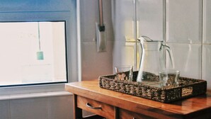 A kitchen counter with a woven tray holding a pitcher and two glasses, a window with a view, and a white tiled wall. - Bajo en la playa con piscina - La Arena 4203 (Arnuero)