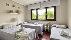 A hotel room with two single beds, a small table with a lamp, a shelf with books and a basket, and a window with a view of greenery.