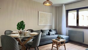 A modern living room with a dining area, a grey sofa, a large window, and a hanging light fixture.