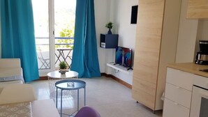 Ein gemütlicher Wohnbereich mit einer Küchenzeile, einem Essbereich mit Tisch und Stühlen, einem kleinen runden Tisch mit Pflanze und einem Balkon mit blauen Vorhängen.
