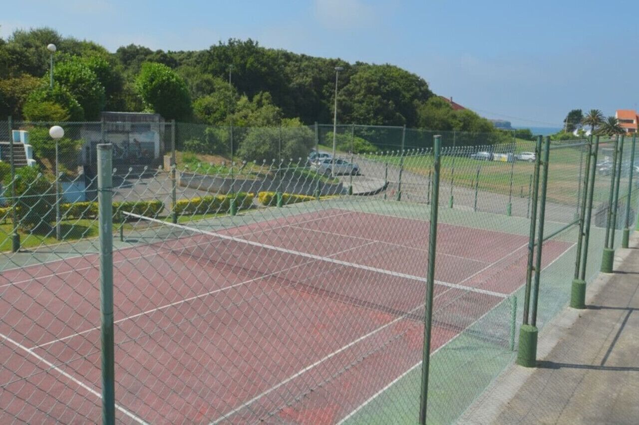 A tennis court enclosed by a chain-link fence with a green net, surrounded by trees and a clear sky.