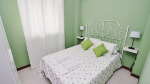 A neatly made bed with white bedding and green pillows, two bedside tables with lamps, and a window with white curtains. - Apartamento con piscina en la playa-La Arena 4501 (Arnuero)