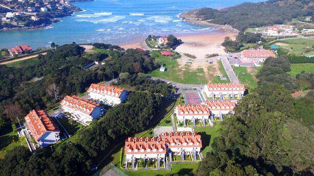 Vista aérea de un complejo turístico costero con varios edificios, una playa y una extensa masa de agua.
