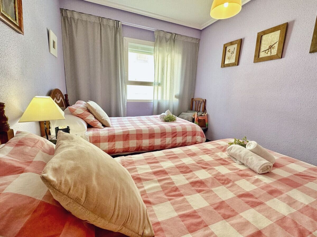 Een hotelkamer met twee bedden, geruit beddengoed, een raam met gordijnen, een lamp en lijstjes met afbeeldingen aan de muur.